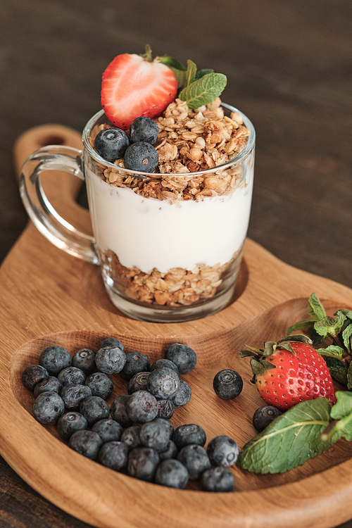 Close-up of glassy mug of granola and summer berries on wooden tray, healthy breakfast in cafe concept