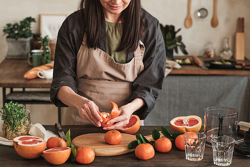 Cropped image of smiling brunette woman in apron standing at table and peeling tangerine above cutting board in kitchen