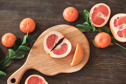 High angle view of two parts of ripe grapefruit on wooden board surrounded citrus fruits and leaves on table