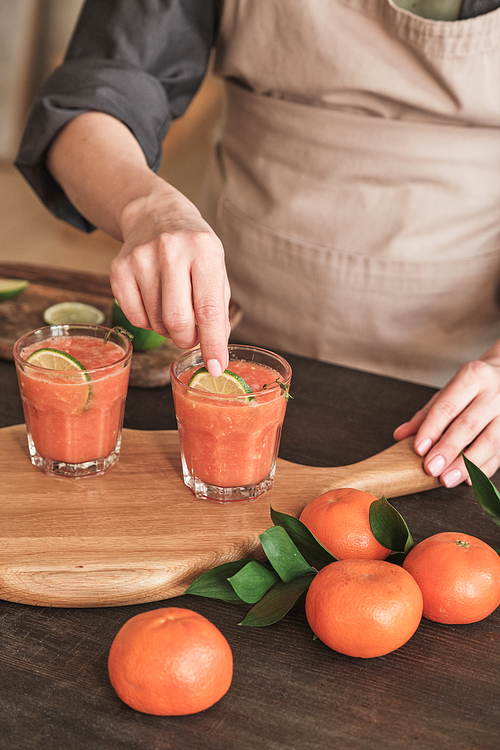 Close-up of unrecognizable woman in apron standing at kitchen counter and decorating tangerine fresh juice with lime slices