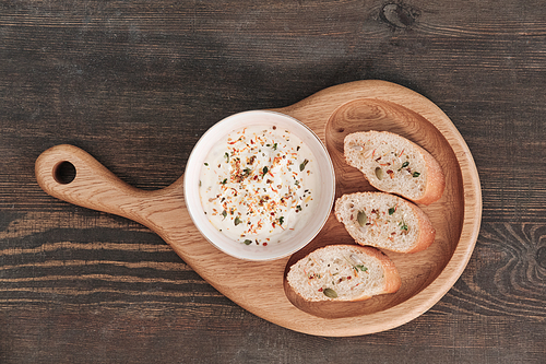Above view of white sauce with spice and slices of baguette on wooden board on dining table