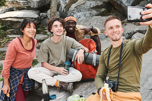 Smiling young man with camera hanging on neck sitting on stone and taking selfie with friends during hike