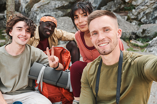 Selfie of smiling young interracial hikers sitting against rocks and posing for camera outdoors