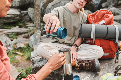 Male hiker sitting with crossed legs on stone and oouring hot water into mug to woman during lunch halt