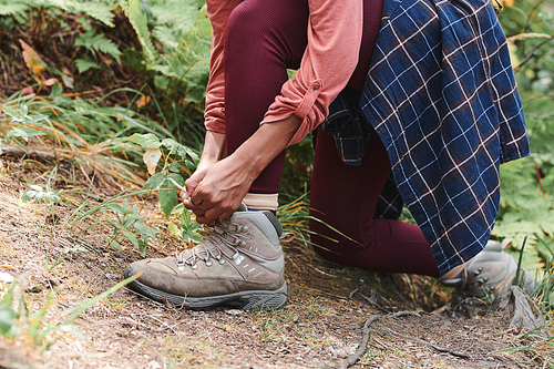 Close-up of unrecognizable woman in maroon leggings standing on one knee and tying boots at hike