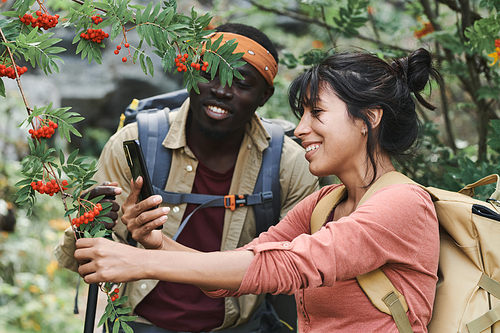 Cheerful young multi-ethnic couple of hikers with backpacks photographing rowanberry in forest