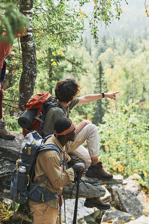 Rear view of young hiker with backpack sitting on stone and showing scenery of forest to black friend