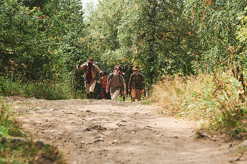 Group of young hikers with bags walking along path in forest in background