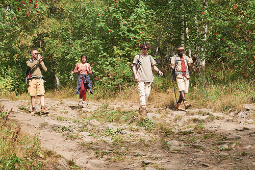 Young man in sunglasses using camera while photographing friends on move at hike in forest