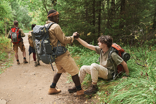 African American hiker with backpack giving hand to fallen man while helping him to stand up during forest hike