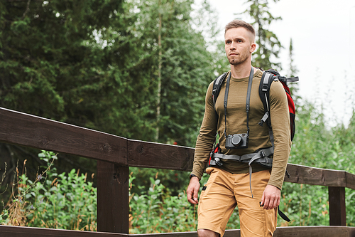 Serious young Caucasian photographer with camera hanging on neck carrying backpack while hiking in forest