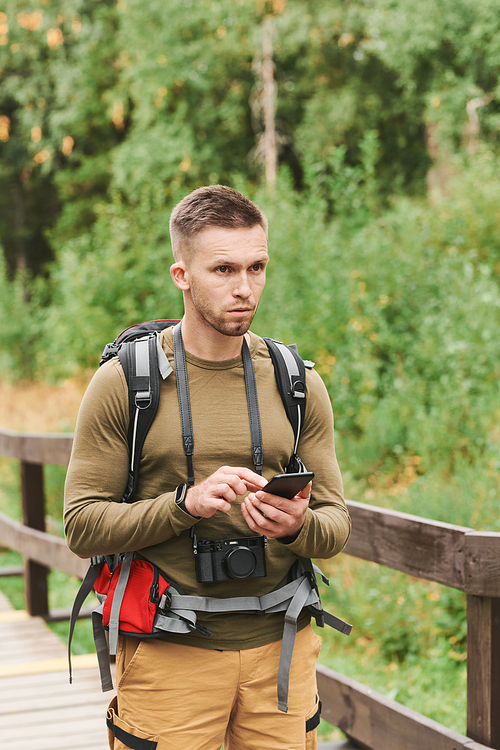 Pensive young hiker with stubble using smartphone while standing on bridge outdoors