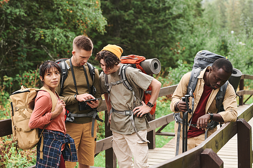 Group of young multi-ethnic hikers with backpacks standing on bridge in forest and analyzing route on phone