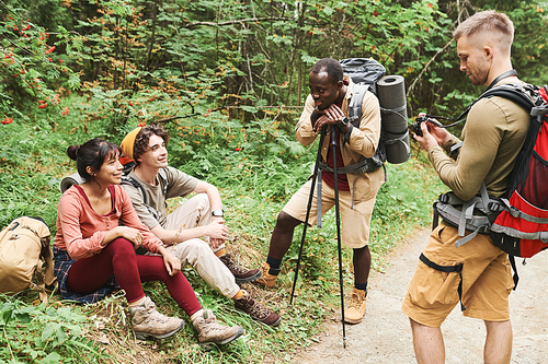 Group of positive young multi-ethnic hikers with backpacks resting on grass and chatting together in forest