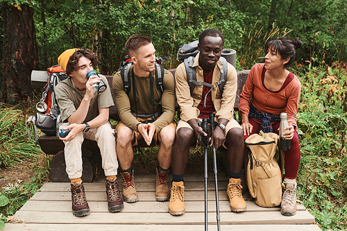 Group of smiling young multi-ethnic friends of hikers sitting on bench in forest and chatting while having break