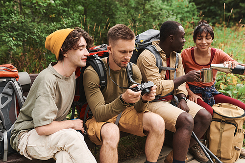 Cheerful young interracial hikers sitting on bench and watching photos and drinking champagne while enjoying adventure together