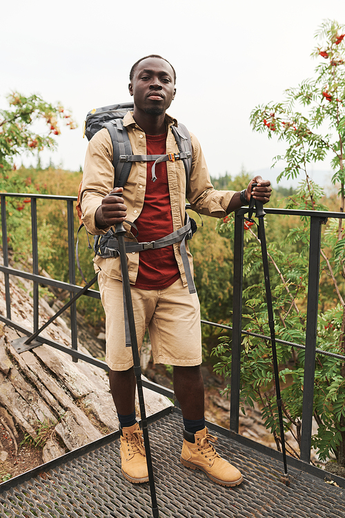 Full length portrait of handsome young Afro-American man with backpack standing with walking poles on bridge outdoors