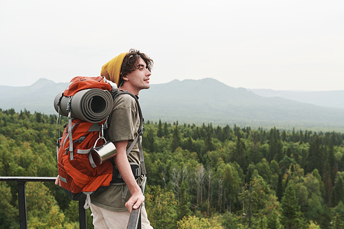 Smiling young Caucasian hiker in yellow hat leaning on railing while contemplating forest