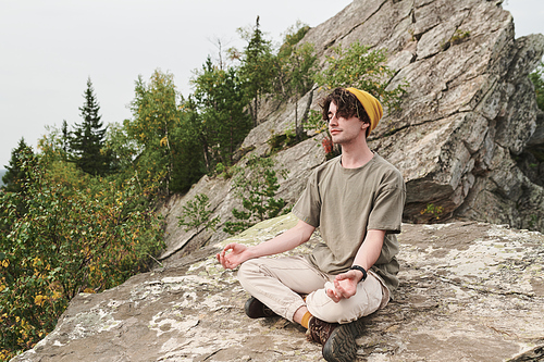 Content relaxed young Caucasian man in hat sitting in lotus position and meditating on big stone outdoors