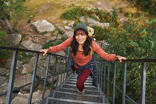 Portrait of positive young mixed race hiker in hat wearing backpack walking up steps in forest