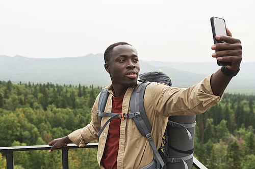 Handsome young Black man with backpack standing on bridge and taking selfie against forest landscape