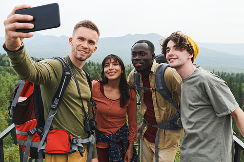 Group of smiling young multi-ethnic hiking friends posing for selfie against forest landscape during adventure