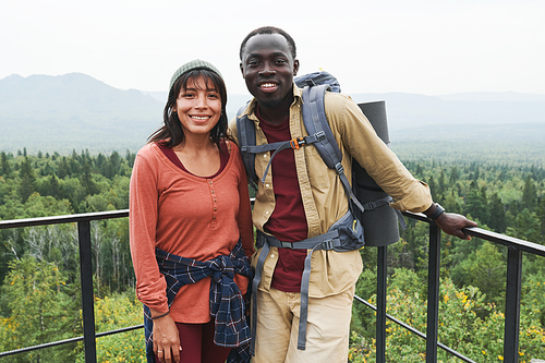 Happy multi-ethnic couple standing at railings against forest landscape and looking at camera at hike