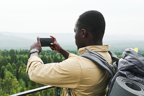 Young African American man with backpack standing on bridge and photographing forest landscape on phone