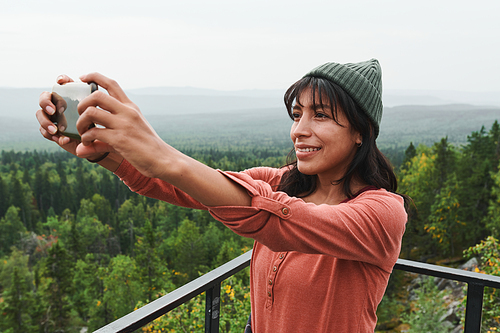 Smiling pretty young mixed race woman in hat standing on bridge and photographing herself on smartphone outdoors