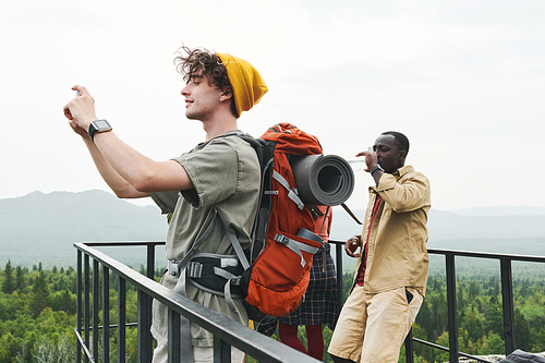 Smiling young hiker with backpack standing on bridge and saving moments while photographing landscape on phone