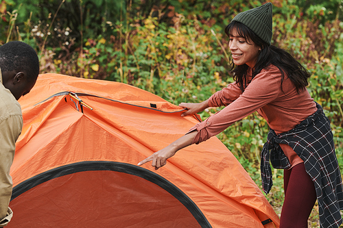 Positive young mixed race woman in hat pointing at pole while assisting boyfriend to set up tent at camping