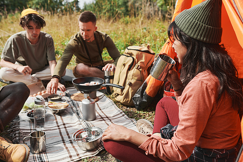 Brunette female hiker in hat sitting with friends at camping in forest and drinking hot tea from metal mug