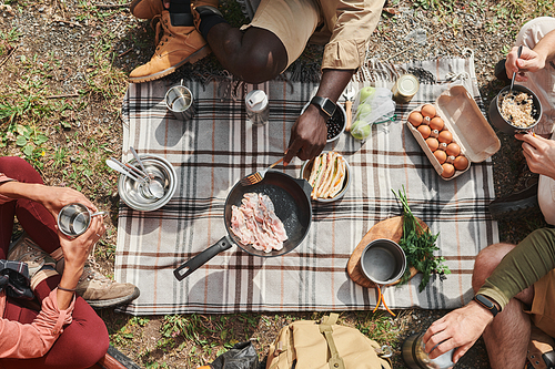 Directly above view of unrecognizable hikers sitting on ground and frying bacon on pan at camping