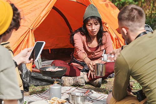 Man pouring water into mug of young woman in hat sitting ar tent outdoors