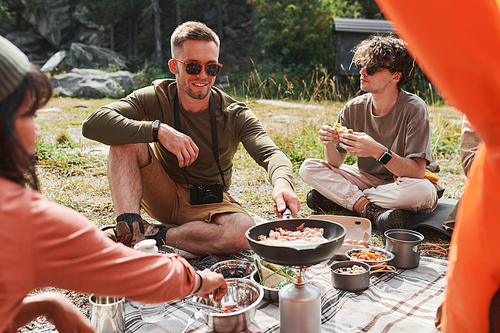 Smiling young Caucasian man in sunglasses sitting among friends and frying eggs on burner at camping