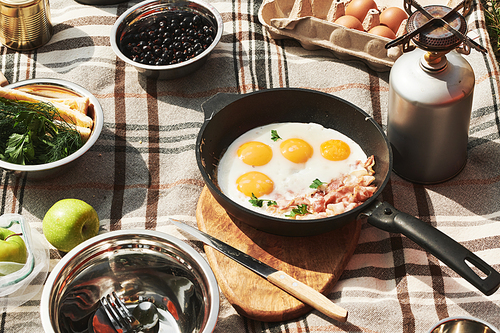 Close-up of fried eggs with bacon on pan and snacks for breakfast outdoors, food on blanket