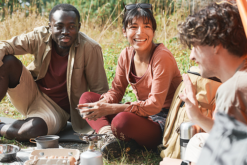 Group of excited young multi-ethnic friends sitting on grass and eating food while resting at forest hike