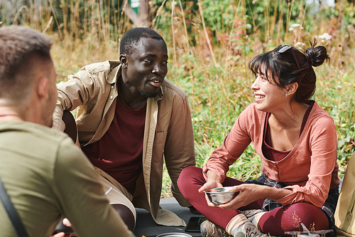 Group of joyful young interracial hikers sitting on ground and chatting while eating during halt