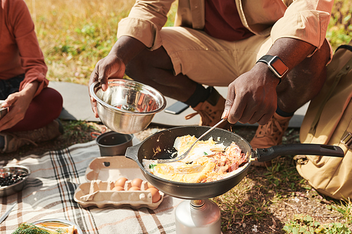 Close-up of unrecognizable black man in smartwatch cooking eggs on burner and putting it in metallic bowl outdoors