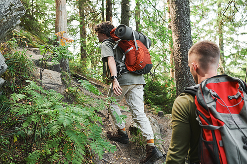 Rear view of young men with backpacks climbing steep cliff while hiking in mountains