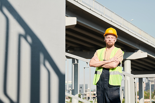 Portrait of confident male construction worker in safety goggles and hardhat standing with crossed arms against bridge