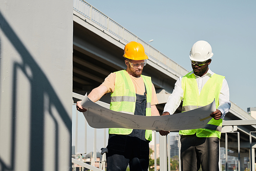 Builder in green vest and hardhat analyzing construction plan with African-American manager at site