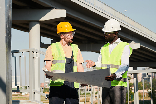 African-American architect in white hardhat and vest pointing at blueprint while working with builder at construction site