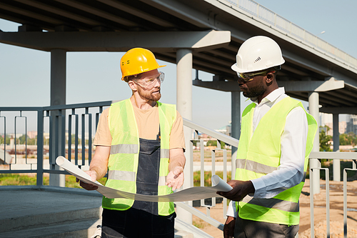 Young African-American construction manager in hardhat and glasses holding blueprint and listening to foreman at building site