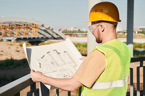 Serious busy man in hardhat and green vest working at construction site of apartment building