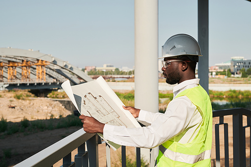 Engineer in hardhat and green vest viewing blueprint and standing on construction site