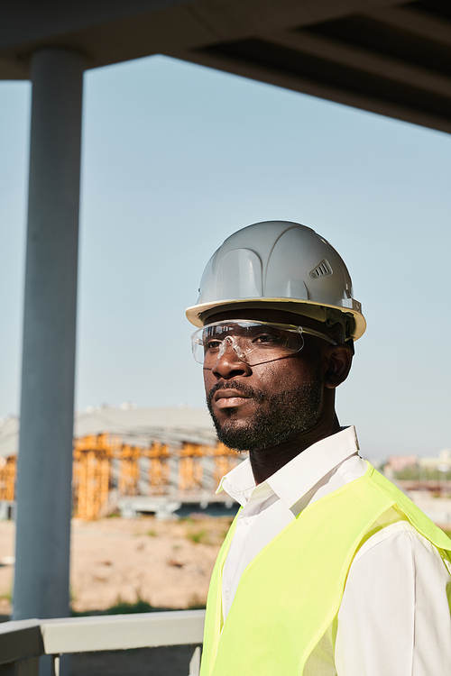 Black Building worker standing on construction site without walls, unfinished block of apartments