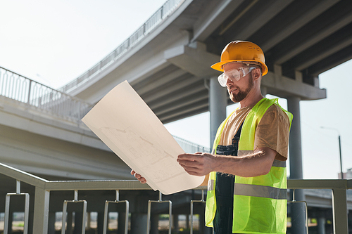 Construction worker examining blueprint and standing on building site of apartment house, large block of flats under construction