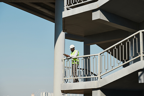 Constriction workers in hardhats and green vests discussing building structure, male engineer pointing at building under