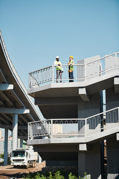 Engineers in white hardhats and safety work vests discussing building project while viewing blueprint at construction site of apartment building